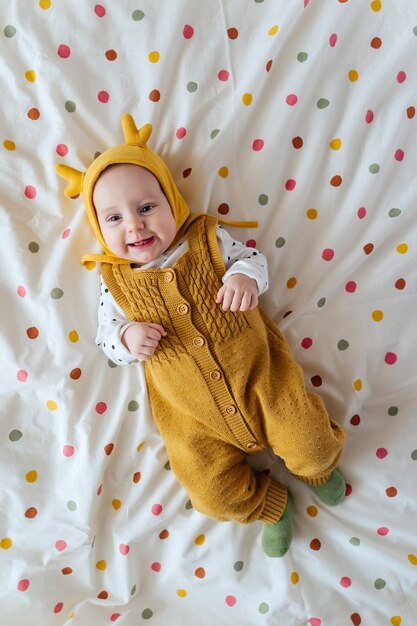 Baby in gebreide stijlvolle kleding en grappige hoed glimlachend en lachend op bed thuis. Mode kindje. Gelukkige jeugd.