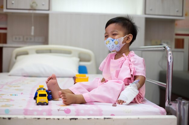 baby in roze patiëntuniform met een masker zit op een bed met een gelukkig gezicht glimlachend in het ziekenhuis