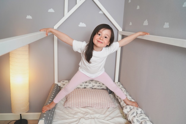 The baby is jumping on his bed before going to bed. a joyful\
girl indulges in her bed. toddler in a white t-shirt and pink\
leggings