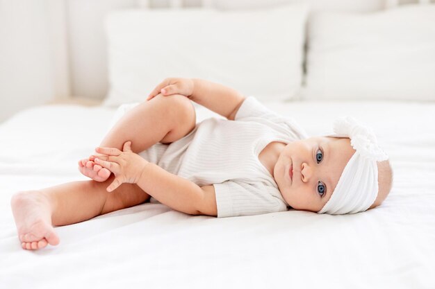 the baby is playing with his legs pushing them into his mouth laughing happy little baby girl with blue eyes in a white bodysuit on a bed in a bright bedroom a newborn baby is smiling happily