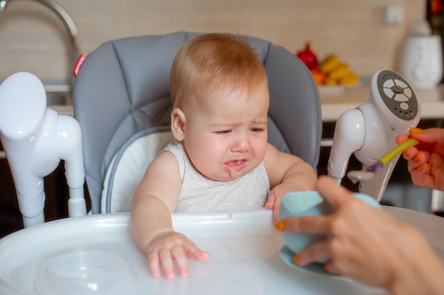 The baby is sitting in a high chair in the kitchen his mother's hand is feeding porridge