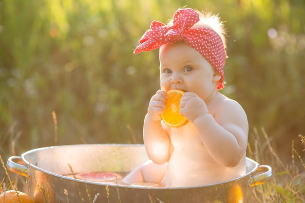 Baby is sitting in an iron tub with oranges and grapefruits in garden and eating a slice of orange