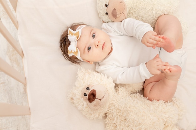 The baby is six months old in a crib in a white bodysuit with a Teddy bear