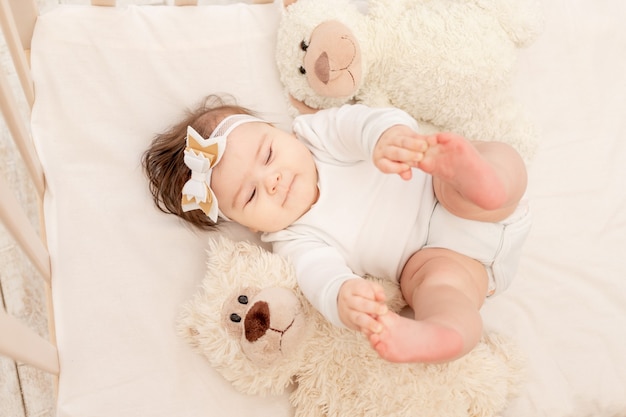 The baby is six months old in a crib in a white bodysuit with a Teddy bear