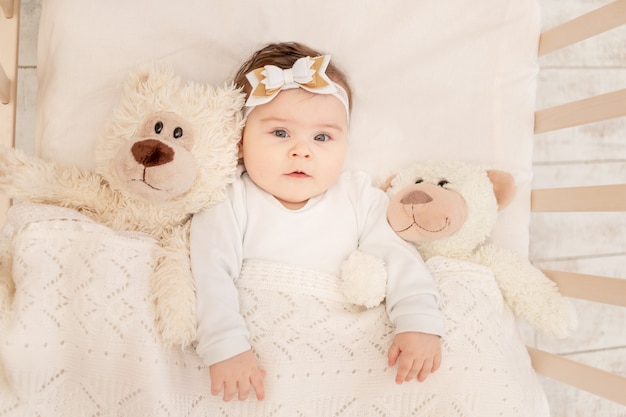 The baby is six months old in a crib in a white bodysuit with a Teddy bear