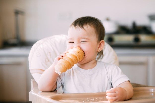 부엌에 있는 아기는 바닐라 크림이 가득한 맛있는 크림 튜브를 탐욕스럽게 먹습니다.