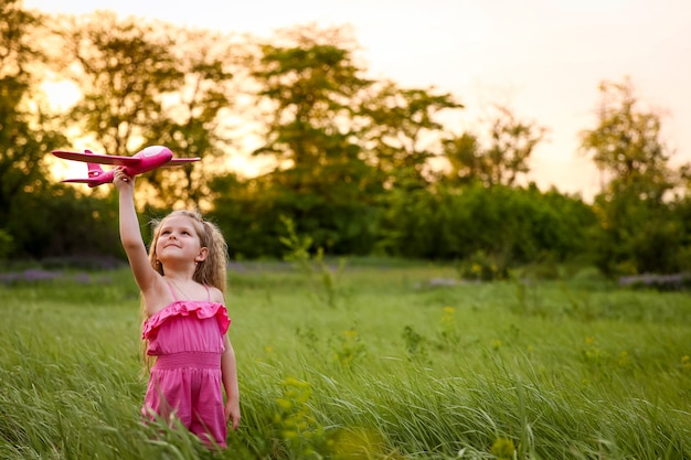 Ребенок запускает розовый самолет на фоне леса и высокой травы. Играя с розовым самолетом в розовом костюме