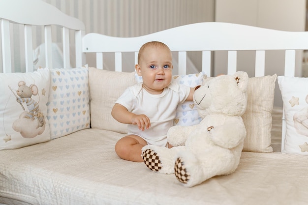 A baby in a light bodysuit in a white crib with a teddy bear toy