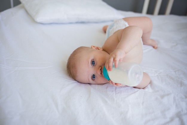 Baby ligt op het bed en drinkt melk uit een fles en kijkt naar de camera