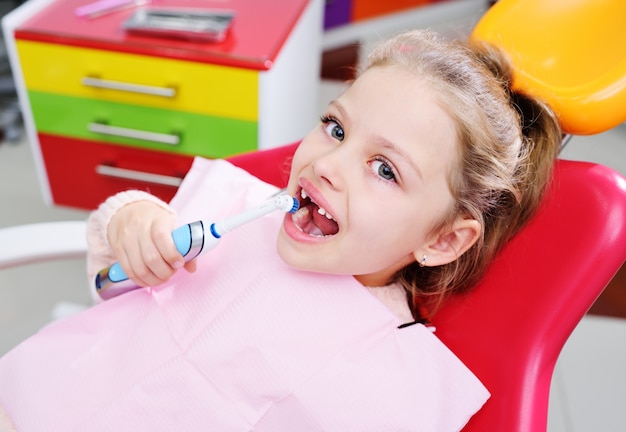 Baby little cute girl without front milk teeth in red dental chair with electric automatic toothbrush in hands.