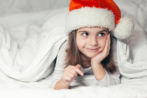 Baby little girl lying in bed in a Santa Claus hat and watching cartoons, studying, playing a game on the phone, looking at the camera, smiling