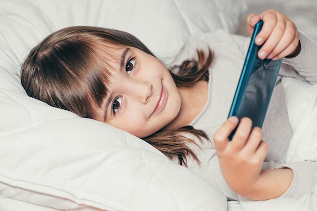 Bambina sdraiata a letto con il telefono e guardando i cartoni animati o giocando al telefono e sorridendo guardando la telecamera
