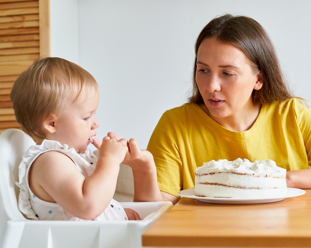 Photo baby looking at creamy cake with interest while sucking finger mother helping daughter eat pie