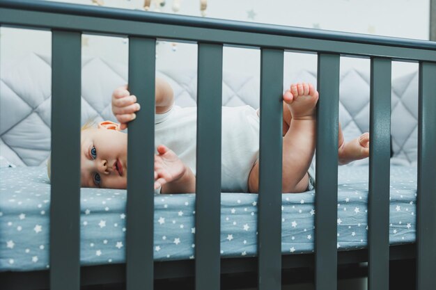 The baby looks through the sides of the crib into the room A small newborn boy lies in a crib for children A place for children to sleep