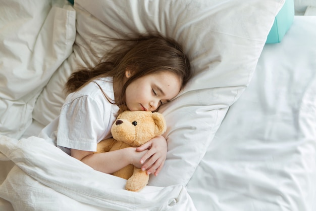 Baby lying in bed with Taddy Bear. Little girl sleeps with knitted toy