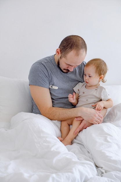 Baby and man sitting on the bed on white linen Toddler looking at father taking temperature