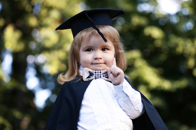 Foto bambino in mantello all'aperto ragazzino bambino in abito accademico nero e cappello scolastico quadrato e farfallino in piedi all'aperto su verde naturale