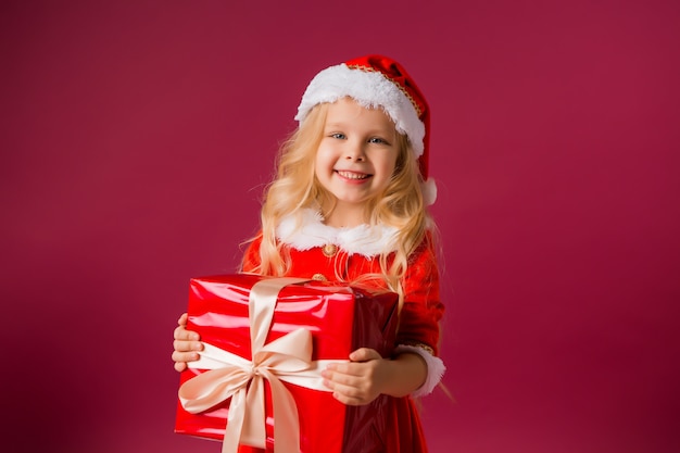 baby meisje in Kerst muts houden geschenkdoos op rode muur, ruimte voor tekst