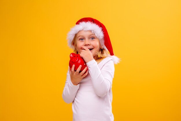 baby meisje in Kerst muts rood spaarvarken op gele muur, ruimte voor tekst te houden