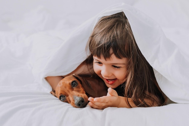 baby meisje knuffelt teckel hond thuis in bed. wit beddengoed. liefde voor huisdieren