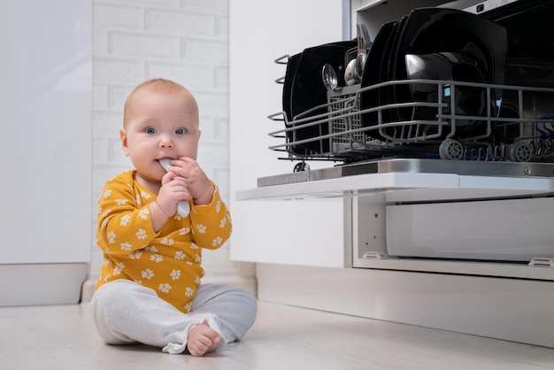 Baby met opgewonden uitdrukking zit op de vloer bij de vaatwasser