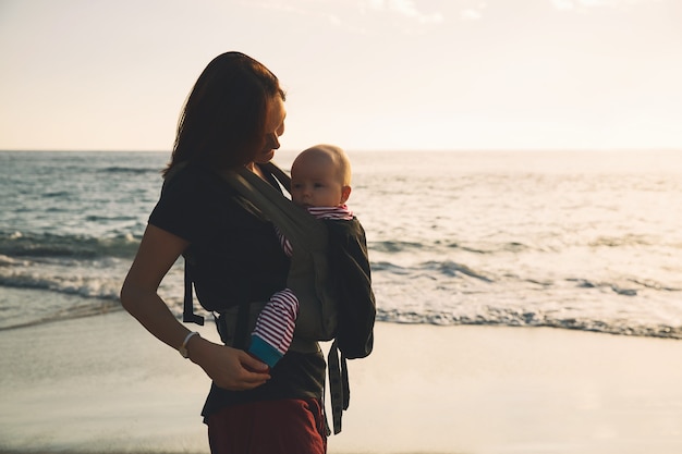 Baby and mother on sea at summer day. Happy family walking on nature outdoors. Child in a carrier backpack. Woman and her baby on coastline ocean on the island of Tenerife, Spain. Travel Europe