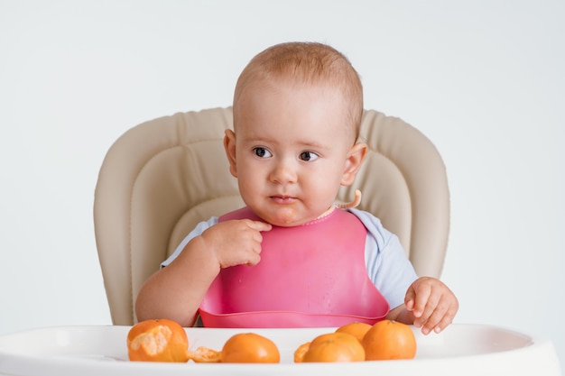 A baby in a pink bib sits on a chair and points a finger questioningly at himself. Lots of tangerines on the table in front of him