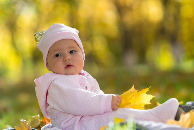 A baby in pink clothing sitting amongst yellow autumn, fall leaves in a park scene.