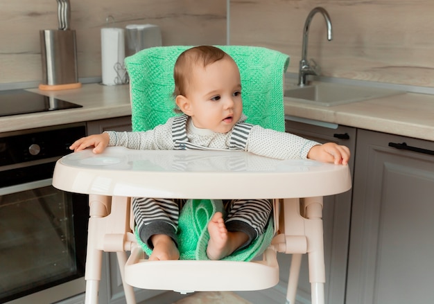 Baby sits in a high chair in the kitchen and eats a banana.