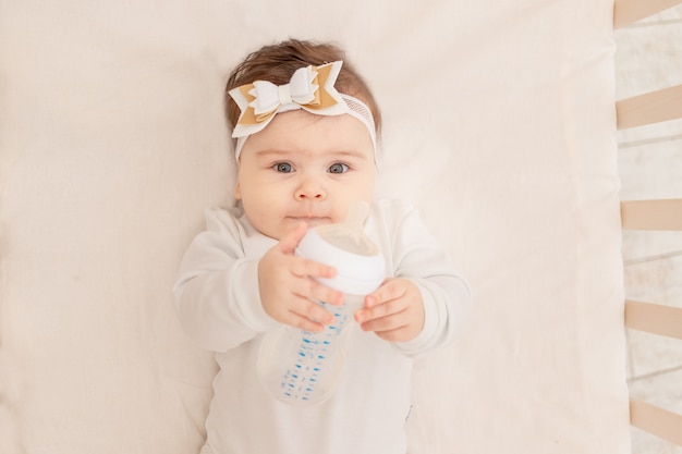 Baby six months lies in a crib with a bottle and drinks water in a white bodysuit