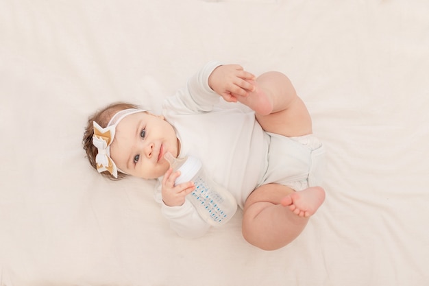 6 개월 아기가 젖병을 들고 침대에 누워 흰색 바디 수트를 입고 물을 마신다