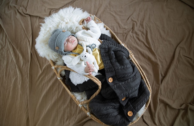 The baby sleeps sweetly in a wicker cradle in a warm knitted hat under a warm blanket with a toy in its handle.