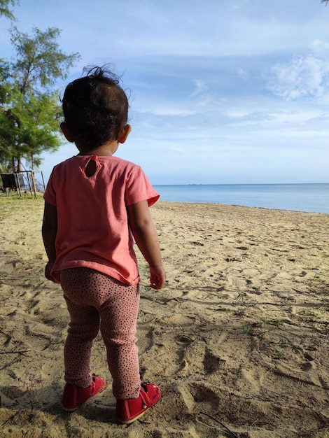 A baby stands on the beach looking at the ocean.