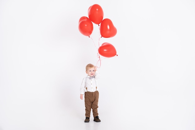 Baby toddler boy in suit holding heart balloon on white wall. Valentine's Day and love concept