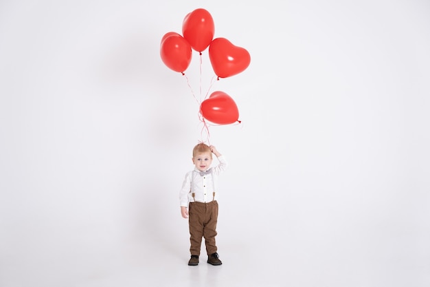 Baby toddler boy in suit holding heart balloon on white.