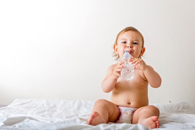 Baby toddler sits on the white bed, smiles and drinks water from plastic bottle