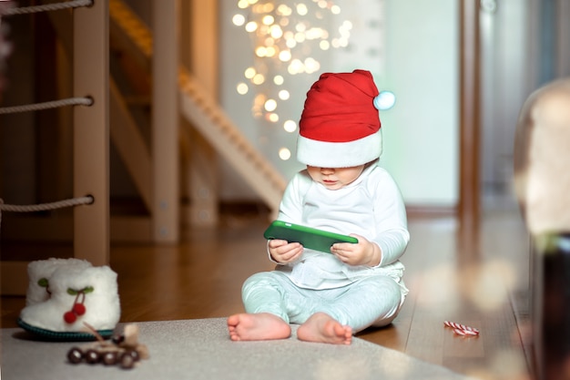 サンタの衣装を着た1歳までの赤ちゃんが、自宅の床に座って興味を持って電話を見ています。