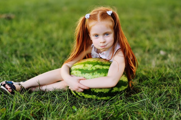 Baby weinig grappig meisje met rood haar die op een reusachtige watermeloen in het Park op het gras op een de zomerdag leunen