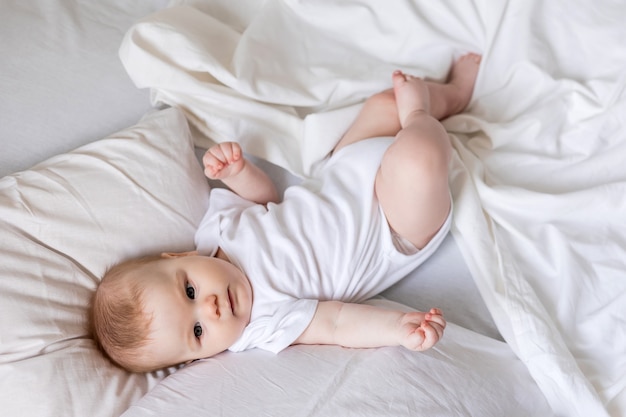 Baby in a white bodysuit is lying on his back in a bed made with white bed linen childrens sleep