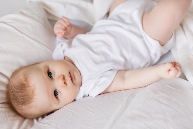 Baby in a white bodysuit is lying on his back in a bed made with white bed linen