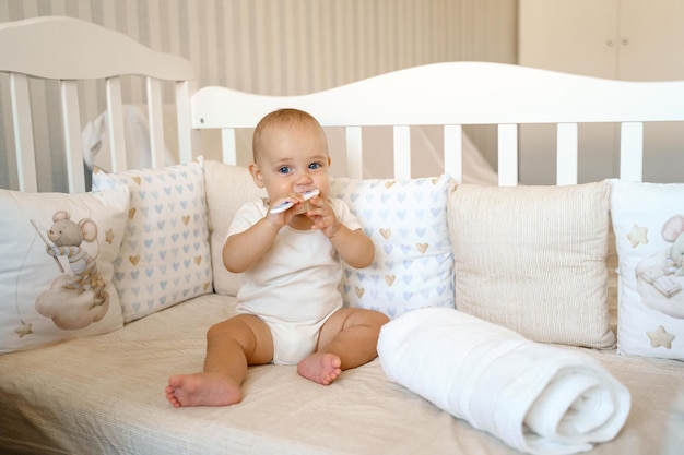 A baby in a white bodysuit is sitting in a crib and gnawing on a comb brush