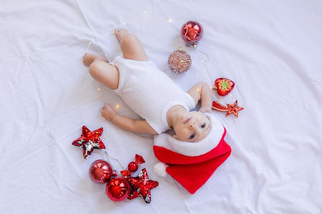 흰색 바디수트와 산타 모자를 쓴 아기가 빨간 크리스마스 트리 장난감으로 둘러싸인 흰색 시트에 등을 대고 누워 있습니다. 겨울, 새해. 텍스트를 위한 공간입니다. 고품질 사진