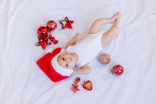 Baby in a white bodysuit and a Santa hat is lying on his back on a white sheet surrounded by red Christmas tree toys. winter, new year. space for text. High quality photo