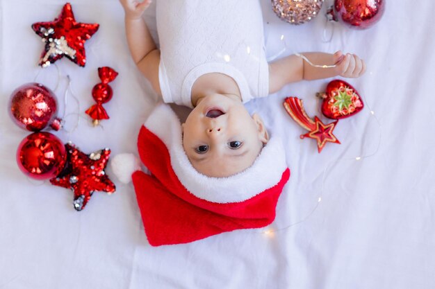 Baby in a white bodysuit and a Santa hat is lying on his back on a white sheet