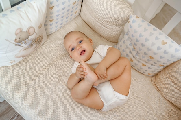 A baby in a white bodysuit in a white crib lies on his back and laughs, holding his legs