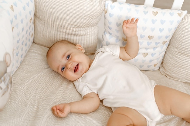 A baby in a white bodysuit in a white crib lies on his back and laughs