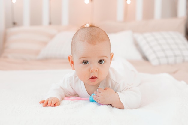 a baby in a white bodysuit with a rattle is lying at home on the bed