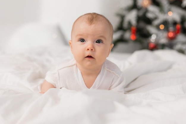 A baby in a white shirt lies on a bed in front of a christmas tree.
