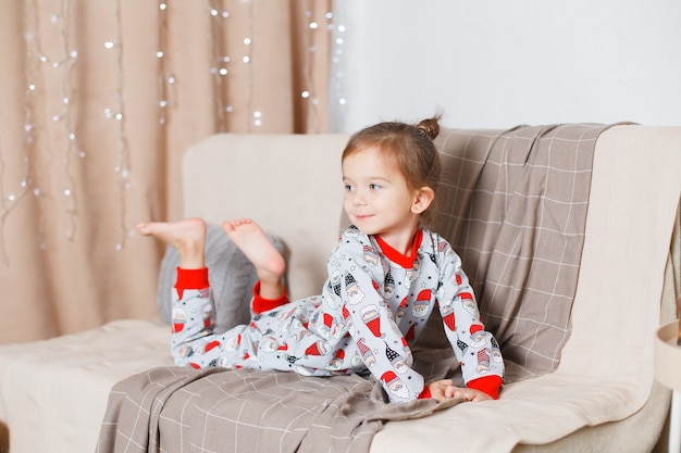 Baby with blonde hair in santa pajamas basics on sofa and daydreaming new Year Christmas