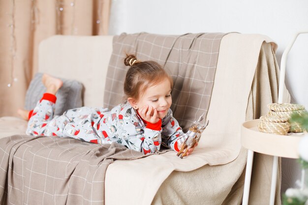 Крошка со светлыми волосами в новогодней пижаме на диване и мечтает о новогоднем Рождестве